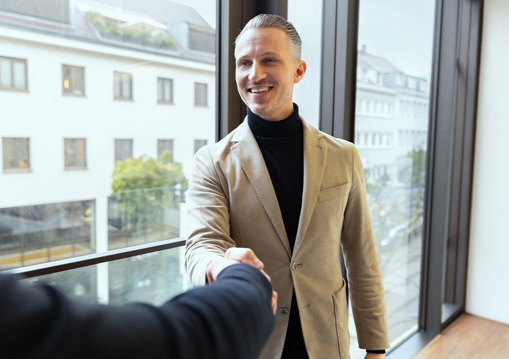 Geschäftsmann im beigen Sakko begrüßt eine Person per Handschlag vor einem Fenster